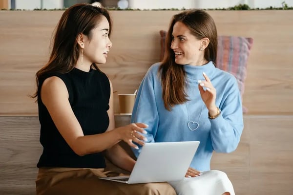 Two female employees sitting down with laptop looking at each other and talking