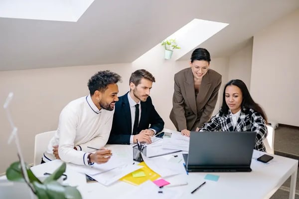 two female employees and two male employees sitting at desk looking into laptop