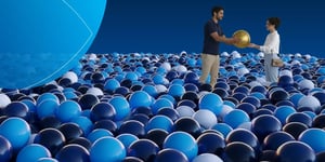Conceptual image of a man and woman standing in a sea of blue and black spheres, handing a single gold sphere to one another.